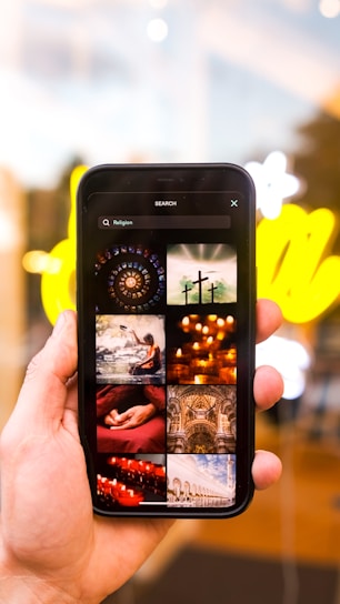 A hand holding a smartphone displaying various images related to religion. The images include crosses, candles, a person meditating, and architectural structures. The background is blurred with bright yellow lights.