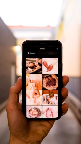 A hand is holding a smartphone displaying images related to newborns. The screen shows a grid of photos, including close-ups of baby feet, a baby wrapped in a blanket, and a baby being held by an adult. The background appears to be an alleyway with blurred walls and a light source visible.