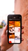 A hand holding a smartphone displaying a search with the keyword 'Seasons'. The phone screen shows a grid of images related to different seasons, such as autumn leaves, a person lying in leaves, a bench, and a snowy train scene. The background is a blurred outdoor setting with a brick wall.