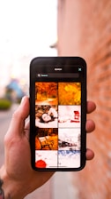 A hand holding a smartphone displaying a search with the keyword 'Seasons'. The phone screen shows a grid of images related to different seasons, such as autumn leaves, a person lying in leaves, a bench, and a snowy train scene. The background is a blurred outdoor setting with a brick wall.