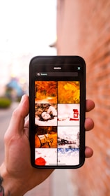A hand holding a smartphone displaying a search with the keyword 'Seasons'. The phone screen shows a grid of images related to different seasons, such as autumn leaves, a person lying in leaves, a bench, and a snowy train scene. The background is a blurred outdoor setting with a brick wall.