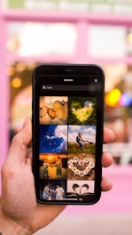 A hand holding a smartphone displaying a search for 'Love' with various images. The background is slightly blurred with a pinkish hue, possibly indicating a storefront or building. The displayed images on the phone include hearts, nature scenes, children, flowers, and books, all conveying themes of affection and warmth.