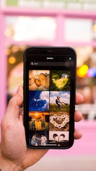 A hand holding a smartphone displaying a search for 'Love' with various images. The background is slightly blurred with a pinkish hue, possibly indicating a storefront or building. The displayed images on the phone include hearts, nature scenes, children, flowers, and books, all conveying themes of affection and warmth.
