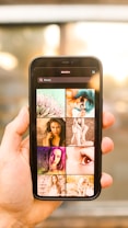 A person holds a smartphone displaying a search screen with the word 'Beauty' and a grid of various images. The images appear to showcase different aspects of beauty, such as portraits of women, close-ups, and artistic visuals. The background is blurred with warm lighting, suggesting an indoor or outdoor setting with sunlight.