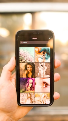 A person holds a smartphone displaying a search screen with the word 'Beauty' and a grid of various images. The images appear to showcase different aspects of beauty, such as portraits of women, close-ups, and artistic visuals. The background is blurred with warm lighting, suggesting an indoor or outdoor setting with sunlight.