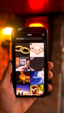 A hand is holding a smartphone displaying a search result for 'wedding' on its screen. The screen shows images related to weddings, such as rings, flowers, and wedding attire. The background features a warm, blurred ambiance with yellow and red lights, hinting at an indoor setting.