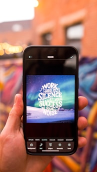A hand holds a smartphone displaying an inspirational quote against a backdrop of a vibrant aurora and a snow-covered landscape. The quote reads 'Work hard in silence, let your success be your noise.' The background features out-of-focus lights and colorful graffiti on a brick wall.