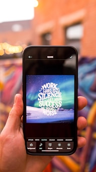 A hand holds a smartphone displaying an inspirational quote against a backdrop of a vibrant aurora and a snow-covered landscape. The quote reads 'Work hard in silence, let your success be your noise.' The background features out-of-focus lights and colorful graffiti on a brick wall.