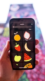 A hand holding a smartphone, which displays a digital drawing app showing colorful cartoon illustrations of various fruits, including an orange, strawberry, banana, apple, watermelon, and others. The background is blurred with colorful, abstract patterns.