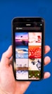 A hand holding a smartphone displaying a search for 'Holiday' with a grid of images. The images include a boat on water at sunset, a red abstract holiday theme, ocean waves, a Christmas tree design, beach chairs, a camera with travel accessories, festive lights, and holiday greenery on a blue background.