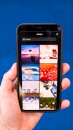 A hand holding a smartphone displaying a search for 'Holiday' with a grid of images. The images include a boat on water at sunset, a red abstract holiday theme, ocean waves, a Christmas tree design, beach chairs, a camera with travel accessories, festive lights, and holiday greenery on a blue background.