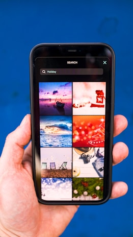 A hand holding a smartphone displaying a search for 'Holiday' with a grid of images. The images include a boat on water at sunset, a red abstract holiday theme, ocean waves, a Christmas tree design, beach chairs, a camera with travel accessories, festive lights, and holiday greenery on a blue background.