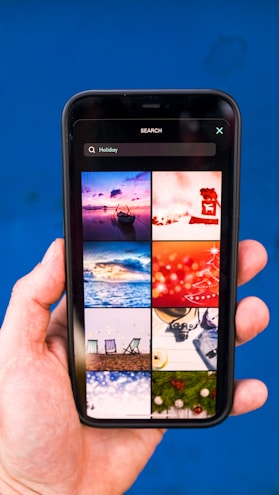 A hand holding a smartphone displaying a search for 'Holiday' with a grid of images. The images include a boat on water at sunset, a red abstract holiday theme, ocean waves, a Christmas tree design, beach chairs, a camera with travel accessories, festive lights, and holiday greenery on a blue background.