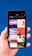 A hand holding a smartphone displaying a search for 'Holiday' with a grid of images. The images include a boat on water at sunset, a red abstract holiday theme, ocean waves, a Christmas tree design, beach chairs, a camera with travel accessories, festive lights, and holiday greenery on a blue background.