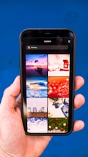 A hand holding a smartphone displaying a search for 'Holiday' with a grid of images. The images include a boat on water at sunset, a red abstract holiday theme, ocean waves, a Christmas tree design, beach chairs, a camera with travel accessories, festive lights, and holiday greenery on a blue background.
