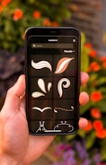 A hand is holding a smartphone displaying a graphic design app, featuring brushstroke and flourish elements against a dark background. In the background, out-of-focus orange flowers and green leaves create a natural setting.