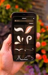 A hand is holding a smartphone displaying a graphic design app, featuring brushstroke and flourish elements against a dark background. In the background, out-of-focus orange flowers and green leaves create a natural setting.