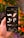 A hand is holding a smartphone displaying a graphic design app, featuring brushstroke and flourish elements against a dark background. In the background, out-of-focus orange flowers and green leaves create a natural setting.