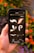 A hand is holding a smartphone displaying a graphic design app, featuring brushstroke and flourish elements against a dark background. In the background, out-of-focus orange flowers and green leaves create a natural setting.