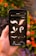 A hand is holding a smartphone displaying a graphic design app, featuring brushstroke and flourish elements against a dark background. In the background, out-of-focus orange flowers and green leaves create a natural setting.