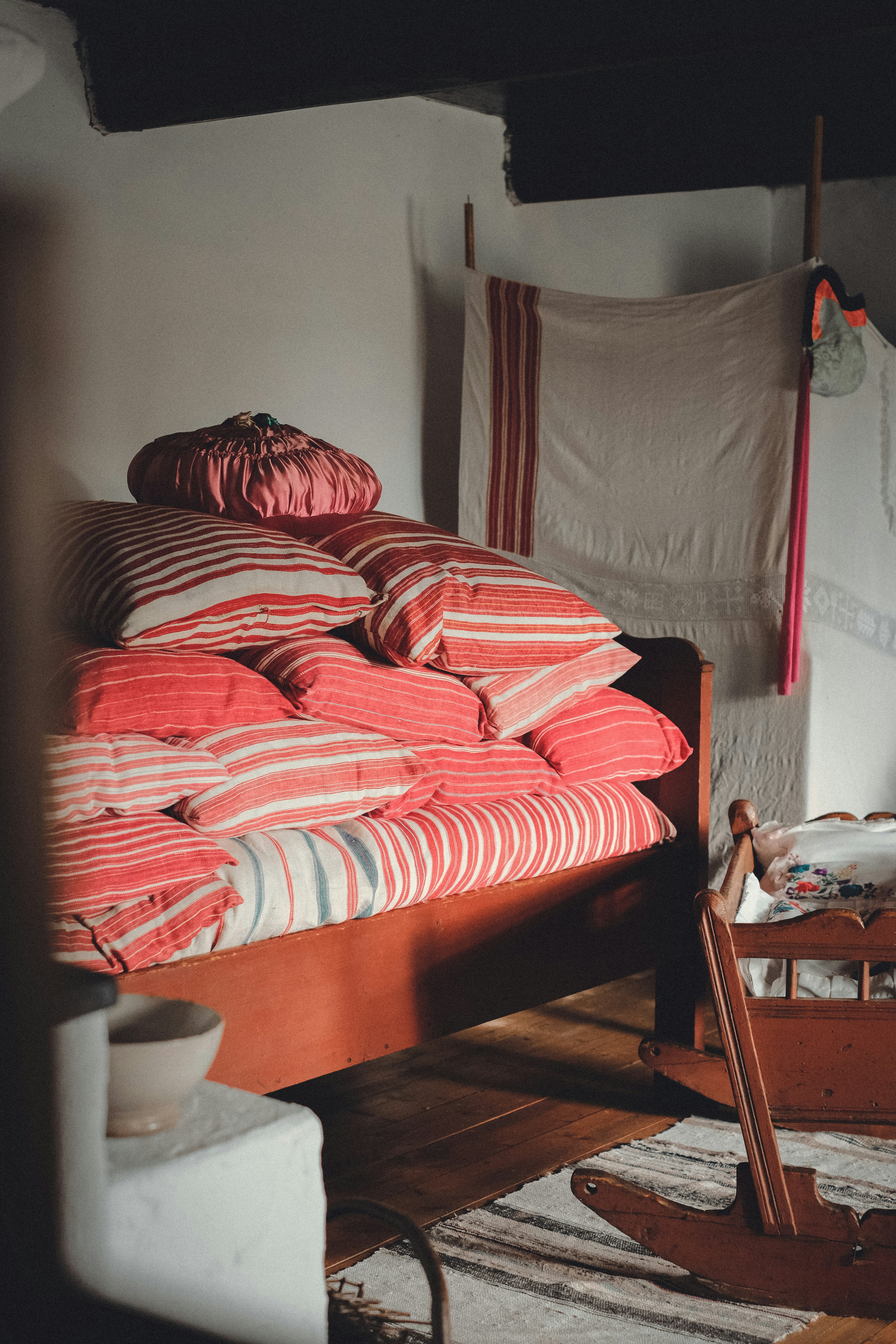 A cozy, rustic room features a wooden bed with a stack of red and striped pillows and blankets. Behind the bed, there is a textile hanging on a rod, displaying a simple pattern. The room includes a wooden cradle or rocking chair on the right side and a white-washed wall partially visible on the left. The warm wooden floor and a small bowl add to the homely ambiance.