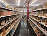 A brightly lit grocery aisle features rows of shelves stocked with a variety of beverage bottles. The selection includes numerous brands of bottled drinks, such as sodas and specialty beverages, displayed in organized sections along both sides of the aisle. The floor is polished and reflective, and the overall ambiance is neat and orderly.