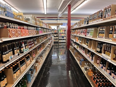 A brightly lit grocery aisle features rows of shelves stocked with a variety of beverage bottles. The selection includes numerous brands of bottled drinks, such as sodas and specialty beverages, displayed in organized sections along both sides of the aisle. The floor is polished and reflective, and the overall ambiance is neat and orderly.