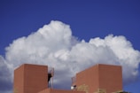 Large white cumulus clouds fill the sky, casting shadows on the vibrant deep blue backdrop. In the foreground, the top of a modern, geometric building with terracotta-hued walls stands prominently. The building features minimalist architectural elements and a small antenna extending upwards.