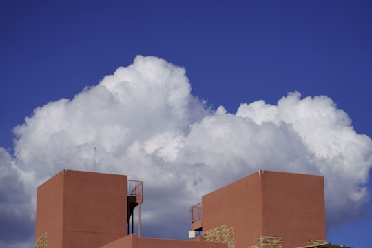 Large white cumulus clouds fill the sky, casting shadows on the vibrant deep blue backdrop. In the foreground, the top of a modern, geometric building with terracotta-hued walls stands prominently. The building features minimalist architectural elements and a small antenna extending upwards.