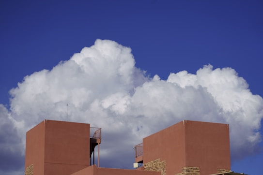 Large white cumulus clouds fill the sky, casting shadows on the vibrant deep blue backdrop. In the foreground, the top of a modern, geometric building with terracotta-hued walls stands prominently. The building features minimalist architectural elements and a small antenna extending upwards.
