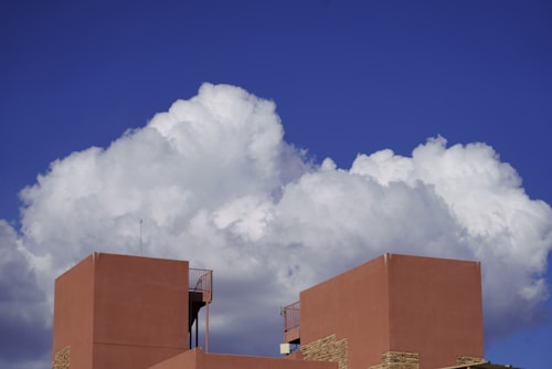 Large white cumulus clouds fill the sky, casting shadows on the vibrant deep blue backdrop. In the foreground, the top of a modern, geometric building with terracotta-hued walls stands prominently. The building features minimalist architectural elements and a small antenna extending upwards.