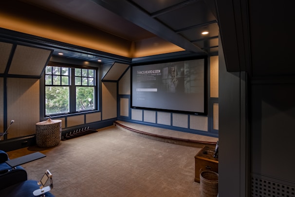 A cozy home theater room features a large projection screen displaying a movie selection interface. The room has dark wood paneling with soft, warm lighting highlighting the ceiling and walls. A large window is situated on one side with an outdoor view partially obscured by trees. There is a curved platform below the screen and a comfortable seating area, including a chevron-patterned ottoman. The room has a neutral color palette with beige, dark gray, and soft brown tones.