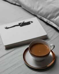 A cup of tea with milk is placed on a white and gold-rimmed saucer. Next to it lies a closed book with a black silhouette of a person on the cover. The setting appears to be on a light-colored bed sheet with a pillow in the background.