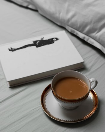 A cup of tea with milk is placed on a white and gold-rimmed saucer. Next to it lies a closed book with a black silhouette of a person on the cover. The setting appears to be on a light-colored bed sheet with a pillow in the background.
