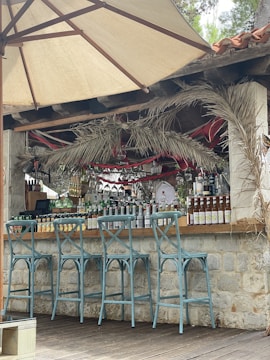 An outdoor bar setup features high chairs painted in light blue, arranged in front of a stone counter. The bar is stocked with various bottles of alcohol, neatly organized behind the counter, and decorated with palm leaves and colorful red ribbons. A large beige umbrella provides shade.
