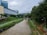 A river flows through an urban area with green pathways on both sides. The scene includes a large building to the left and dense trees along the right path. Overcast skies add to the mood, and there is a small bridge or crossing visible in the distance.