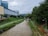 A river flows through an urban area with green pathways on both sides. The scene includes a large building to the left and dense trees along the right path. Overcast skies add to the mood, and there is a small bridge or crossing visible in the distance.