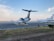 Several private jets are parked on a tarmac at an airport under a blue sky with scattered clouds. The ground is lined with grassy patches and a few traffic cones. An aircraft is taking off or landing in the background.