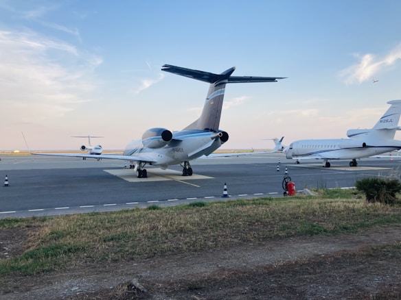 Several private jets are parked on a tarmac at an airport under a blue sky with scattered clouds. The ground is lined with grassy patches and a few traffic cones. An aircraft is taking off or landing in the background.