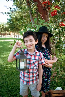 A boy and girl are standing outdoors in a garden. The boy is holding a lantern and wearing a checkered shirt and shorts. The girl is wearing a dress with floral patterns and a black wide-brimmed hat. They are surrounded by greenery and red flowers, with a large tree in the background.