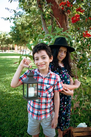 A boy and girl are standing outdoors in a garden. The boy is holding a lantern and wearing a checkered shirt and shorts. The girl is wearing a dress with floral patterns and a black wide-brimmed hat. They are surrounded by greenery and red flowers, with a large tree in the background.