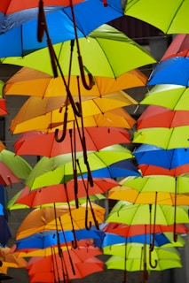 Multiple colorful umbrellas hanging upside down in rows create a vibrant canopy. The umbrellas are predominantly red, blue, yellow, green, and orange, with wooden handles visible underneath.