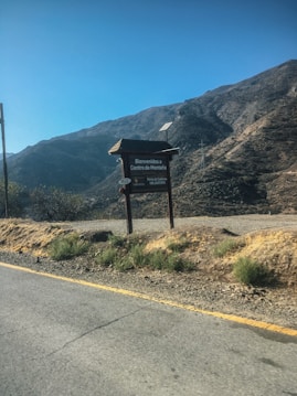 A mountainous landscape with dry terrain and sparse vegetation under a clear blue sky. There is a road with a visible yellow line on the side, and a sign next to the road that reads 'Bienvenidos a Centro de Monta&ntilde;a' indicating a mountain center. The surrounding hills are mostly barren with patches of shrubs and rocks.