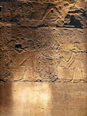 Carved into a stone surface are ancient Egyptian-style hieroglyphs and figures. The detailed engraving features two human-like figures in traditional attire, likely engaged in a ritual or ceremony. Intricate patterns and symbols surround them, suggesting a scene of cultural or historical significance.