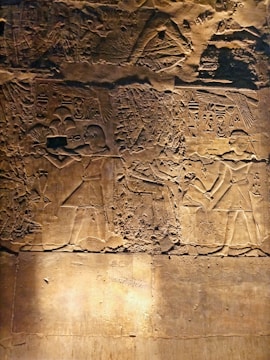 Carved into a stone surface are ancient Egyptian-style hieroglyphs and figures. The detailed engraving features two human-like figures in traditional attire, likely engaged in a ritual or ceremony. Intricate patterns and symbols surround them, suggesting a scene of cultural or historical significance.