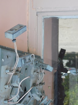 A close-up view of an old electrical panel with multiple metallic boxes and wires. The boxes are mounted on a gray metal surface, and the wires appear tangled and unorganized. There is a dirty glass window on the right side, reflecting some outdoor elements.