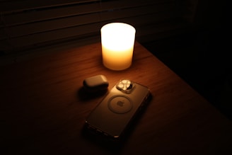 A smartphone and wireless earbud case rest on a wooden surface, softly illuminated by the warm glow of a lit candle.