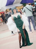 A person with blue hair, wearing sunglasses, a green strappy dress, and a white fur shawl is standing confidently at an outdoor event. The individual accessorizes with layered pearl necklaces and wears black lace stockings and strappy high heels. The background features several people walking and tents, indicating a lively atmosphere.