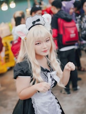 A person dressed in a maid costume with cat ears, featuring long blonde hair and a playful expression. They are surrounded by a crowd of people, some wearing backpacks.