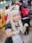 A person dressed in a maid costume with cat ears, featuring long blonde hair and a playful expression. They are surrounded by a crowd of people, some wearing backpacks.